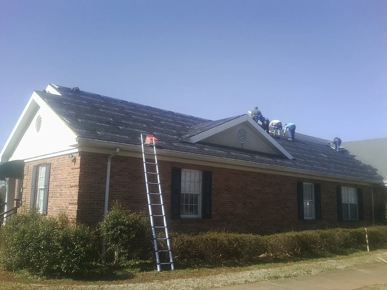 Crew tearing shingles off a brick ranch roof with a ladder set — Metal Roof Repair work by our crew in Spring Grove
