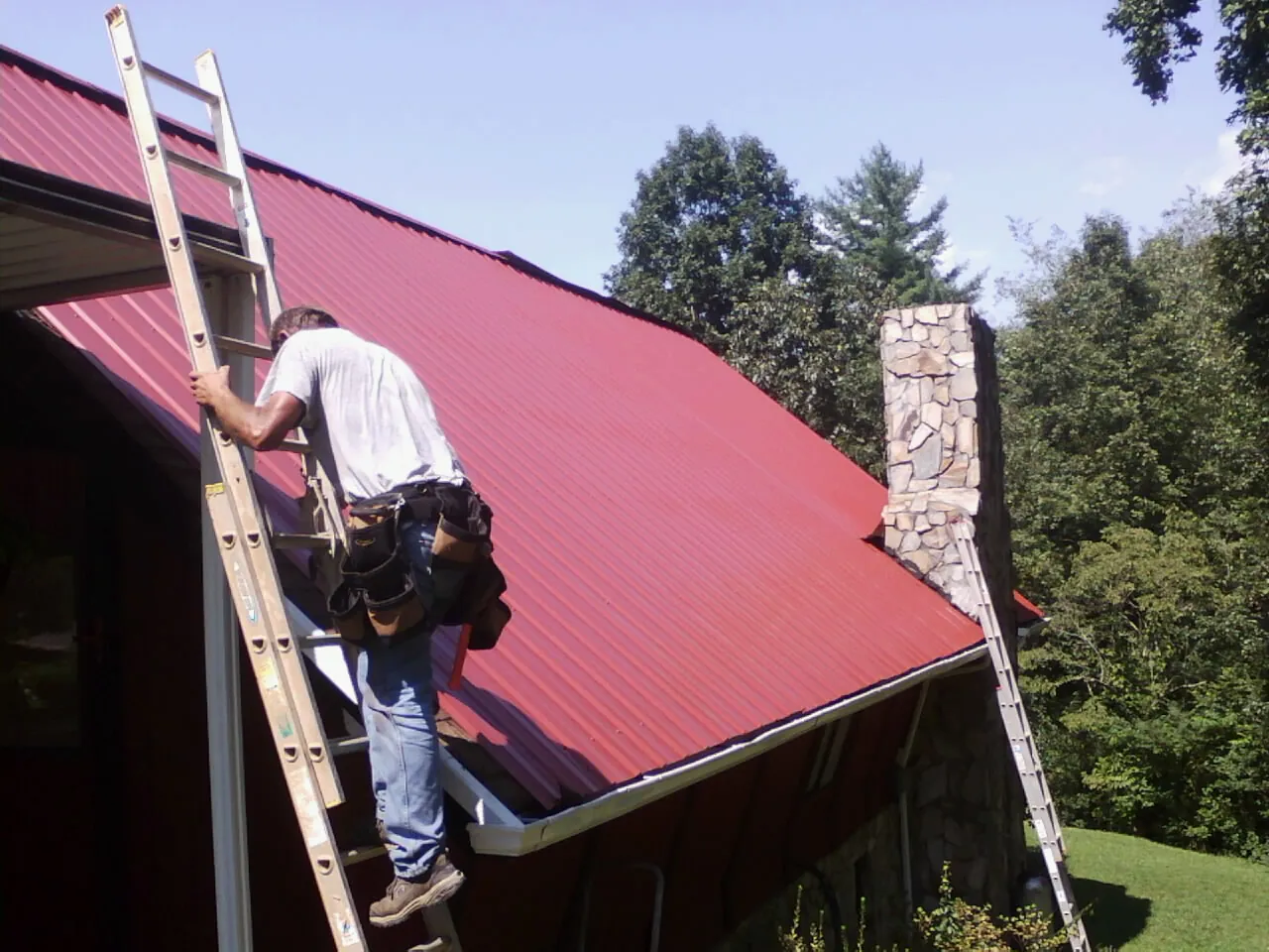 Roofer on a ladder beside a red metal roof and stone chimney — Soffit & Fascia Repair by Roofing Brothers in Spring Grove